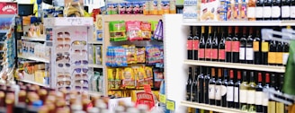 A grocery store aisle with a display of sunglasses, various packaged snacks, and shelves stocked with multiple bottles of wine. The foreground shows a rack with colorful packaging and bright labels, while the background contains an assortment of alcoholic beverages.