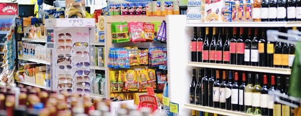 A grocery store aisle with a display of sunglasses, various packaged snacks, and shelves stocked with multiple bottles of wine. The foreground shows a rack with colorful packaging and bright labels, while the background contains an assortment of alcoholic beverages.