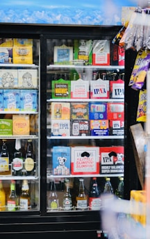 A refrigerated display case filled with a variety of alcoholic beverages, including craft beers and some popular brands. The top row displays brightly colored packaging with different logos, while the bottom shelves contain various bottled drinks arranged neatly. To the right, some snack items hang on the outside of the case.