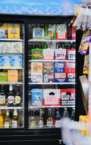 A refrigerated display case filled with a variety of alcoholic beverages, including craft beers and some popular brands. The top row displays brightly colored packaging with different logos, while the bottom shelves contain various bottled drinks arranged neatly. To the right, some snack items hang on the outside of the case.