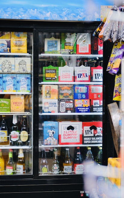 A refrigerated display case filled with a variety of alcoholic beverages, including craft beers and some popular brands. The top row displays brightly colored packaging with different logos, while the bottom shelves contain various bottled drinks arranged neatly. To the right, some snack items hang on the outside of the case.