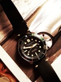 A wristwatch with a black leather strap and a dark dial is placed on top of a magazine or catalog featuring a car image. The watch has bold hour markers and numerals on the bezel, with what appears to be hands displaying the time.