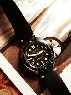 A wristwatch with a black leather strap and a dark dial is placed on top of a magazine or catalog featuring a car image. The watch has bold hour markers and numerals on the bezel, with what appears to be hands displaying the time.