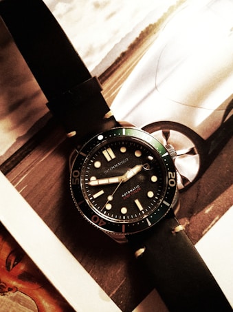 A wristwatch with a black leather strap and a dark dial is placed on top of a magazine or catalog featuring a car image. The watch has bold hour markers and numerals on the bezel, with what appears to be hands displaying the time.