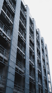 A modern, industrial-style building featuring a repetitive pattern of metal panels and balconies. The structure is composed of steel framework and numerous windows with a sleek, geometric design. The sky in the background is overcast, providing a stark contrast to the dark, metallic façade.