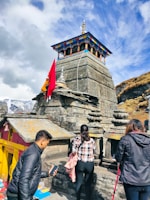 A stone temple with colorful decorations at the top stands against a backdrop of cloudy skies and mountains. People are seen near the temple, some holding phones and others standing close to the structure. A red flag is prominently displayed attached to the temple, adding vibrancy to the scene.