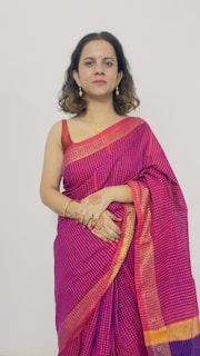 A woman wearing a traditional pink and purple sari with gold accents stands against a plain background. Her hands are adorned with intricate henna designs, and she is wearing gold jewelry including earrings and a necklace.
