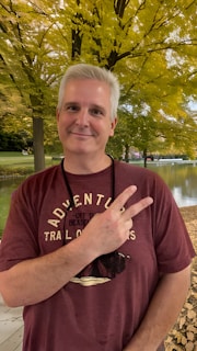 A person with light hair is smiling and holding up a peace sign with one hand. They are wearing a maroon T-shirt with text and graphics. The background features a serene outdoor setting with lush green and yellow leaves, indicating a park or natural setting.
