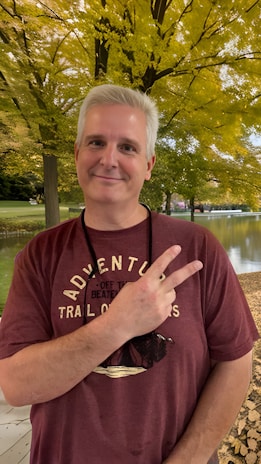 A person with light hair is smiling and holding up a peace sign with one hand. They are wearing a maroon T-shirt with text and graphics. The background features a serene outdoor setting with lush green and yellow leaves, indicating a park or natural setting.