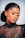 A person with elegant makeup and neatly styled hair is wearing a black, sequined, sheer top. The makeup features bold, colorful eyeshadow and long, dramatic eyelashes. The expression is serene and the pose is slightly tilted, highlighting the intricate details of the outfit.