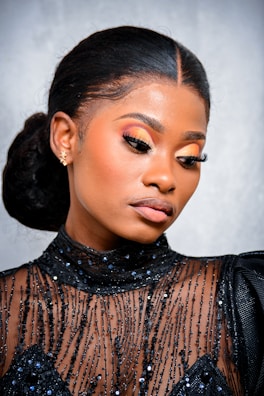 A person with elegant makeup and neatly styled hair is wearing a black, sequined, sheer top. The makeup features bold, colorful eyeshadow and long, dramatic eyelashes. The expression is serene and the pose is slightly tilted, highlighting the intricate details of the outfit.