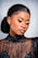 A person with elegant makeup and neatly styled hair is wearing a black, sequined, sheer top. The makeup features bold, colorful eyeshadow and long, dramatic eyelashes. The expression is serene and the pose is slightly tilted, highlighting the intricate details of the outfit.