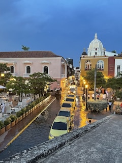 A vibrant street scene during dusk with pastel-colored colonial buildings and ornate architecture. Yellow taxis line the street, reflecting lights on the wet cobblestone. People stroll along the sidewalk on a lively evening in an urban setting.