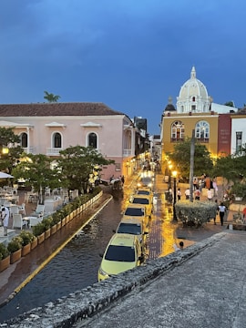 A vibrant street scene during dusk with pastel-colored colonial buildings and ornate architecture. Yellow taxis line the street, reflecting lights on the wet cobblestone. People stroll along the sidewalk on a lively evening in an urban setting.