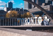 Large white letters spelling 'Toronto' stand prominently amidst an urban setting. Behind the sign, there are modern high-rise buildings and trees with autumn foliage. A person with a colorful backpack is seated in the foreground near a metal railing. The sky is clear with some clouds, adding to the lively cityscape atmosphere.