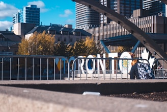 Large white letters spelling 'Toronto' stand prominently amidst an urban setting. Behind the sign, there are modern high-rise buildings and trees with autumn foliage. A person with a colorful backpack is seated in the foreground near a metal railing. The sky is clear with some clouds, adding to the lively cityscape atmosphere.