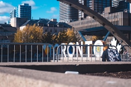 Large white letters spelling 'Toronto' stand prominently amidst an urban setting. Behind the sign, there are modern high-rise buildings and trees with autumn foliage. A person with a colorful backpack is seated in the foreground near a metal railing. The sky is clear with some clouds, adding to the lively cityscape atmosphere.