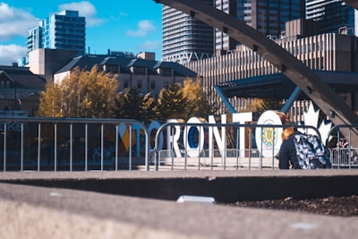 Large white letters spelling 'Toronto' stand prominently amidst an urban setting. Behind the sign, there are modern high-rise buildings and trees with autumn foliage. A person with a colorful backpack is seated in the foreground near a metal railing. The sky is clear with some clouds, adding to the lively cityscape atmosphere.