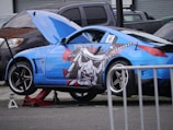 A blue sports car is elevated on a jack, with its front hood open for maintenance. The car features a bold graphic on its side, depicting a samurai helmet in black, gray, and red. Other vehicles are parked nearby.