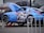 A blue sports car is elevated on a jack, with its front hood open for maintenance. The car features a bold graphic on its side, depicting a samurai helmet in black, gray, and red. Other vehicles are parked nearby.