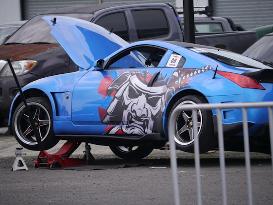 A blue sports car is elevated on a jack, with its front hood open for maintenance. The car features a bold graphic on its side, depicting a samurai helmet in black, gray, and red. Other vehicles are parked nearby.
