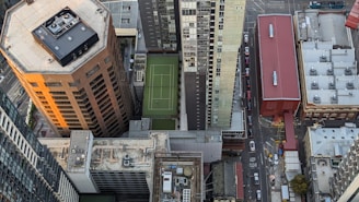 Aerial view of an urban area with multiple high-rise buildings. Prominent among them is a tall, octagonal building with a beige facade and a flat rooftop equipped with machinery. Adjacent to it is a rectangular residential skyscraper with various balconies. In between the buildings, a green rooftop sports court is visible. The scene includes a narrow street lined with vehicles, alongside a red-roofed industrial structure.