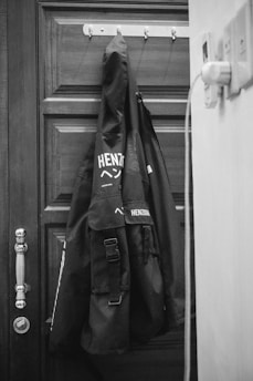 A black coat is hanging on a hook attached to a wooden door. There is a visible logo with the text that appears to say 'Henzorg' on the coat. The door has a classic design with a metallic handle, and a piece of electrical equipment is plugged into a socket nearby.