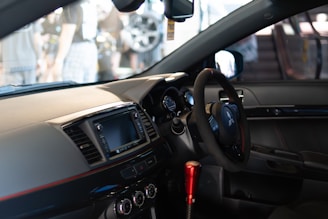 A modern car interior featuring a steering wheel with the Mitsubishi logo, a digital display screen, and several control buttons and dials on the dashboard. The gear shift is prominently visible with a metallic red finish, and the seating is upholstered in dark leather.