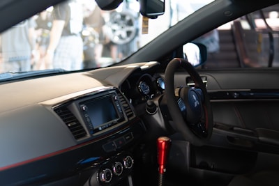 A modern car interior featuring a steering wheel with the Mitsubishi logo, a digital display screen, and several control buttons and dials on the dashboard. The gear shift is prominently visible with a metallic red finish, and the seating is upholstered in dark leather.