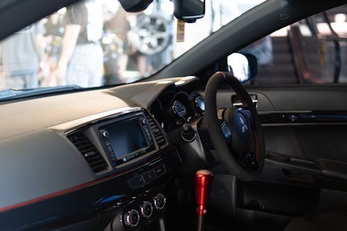 A modern car interior featuring a steering wheel with the Mitsubishi logo, a digital display screen, and several control buttons and dials on the dashboard. The gear shift is prominently visible with a metallic red finish, and the seating is upholstered in dark leather.