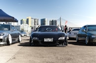 A car meet takes place in an urban setting with multiple modified cars parked on a wide pavement. A shiny black sports car with a 'DEPORT' license plate is centered among other vehicles. High-rise buildings and a cable-stayed bridge form the backdrop. A blue and white canopy tent is set up on the left, while small groups of people are scattered around the area, suggesting a casual gathering or event.