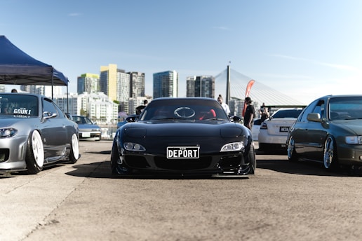 A car meet takes place in an urban setting with multiple modified cars parked on a wide pavement. A shiny black sports car with a 'DEPORT' license plate is centered among other vehicles. High-rise buildings and a cable-stayed bridge form the backdrop. A blue and white canopy tent is set up on the left, while small groups of people are scattered around the area, suggesting a casual gathering or event.