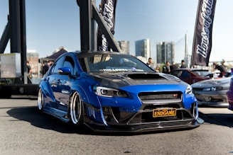 A modified blue sports car with a sleek design is prominently placed in the foreground of an outdoor setting, likely a car show. The car has a custom black grille and a license plate that reads 'ENDGAME'. Background elements include banners, another car, people milling around, a cityscape with tall buildings, and a bridge structure, highlighting a bustling urban environment.
