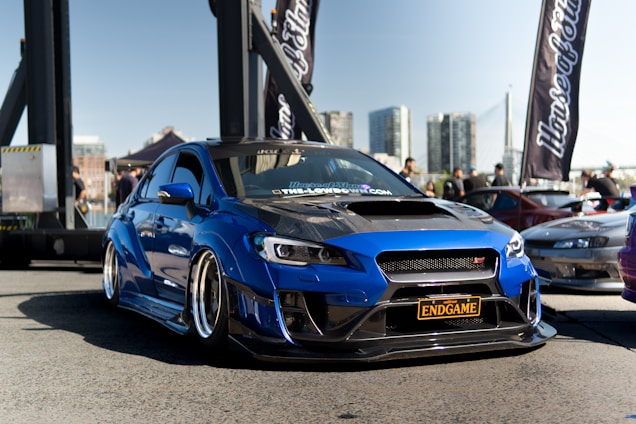 A modified blue sports car with a sleek design is prominently placed in the foreground of an outdoor setting, likely a car show. The car has a custom black grille and a license plate that reads 'ENDGAME'. Background elements include banners, another car, people milling around, a cityscape with tall buildings, and a bridge structure, highlighting a bustling urban environment.