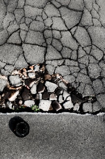 Cracked asphalt pavement with visible underlying stones and gravel. A small patch of green vegetation emerges through the cracks. The surface has a rough, uneven texture with varying shades of gray.