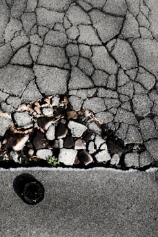Cracked asphalt pavement with visible underlying stones and gravel. A small patch of green vegetation emerges through the cracks. The surface has a rough, uneven texture with varying shades of gray.