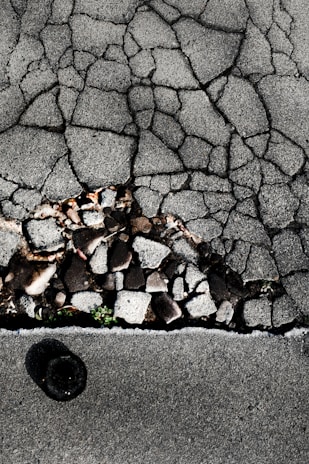Cracked asphalt pavement with visible underlying stones and gravel. A small patch of green vegetation emerges through the cracks. The surface has a rough, uneven texture with varying shades of gray.