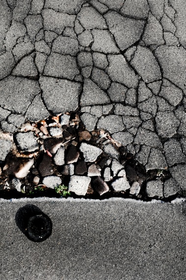 Cracked asphalt pavement with visible underlying stones and gravel. A small patch of green vegetation emerges through the cracks. The surface has a rough, uneven texture with varying shades of gray.