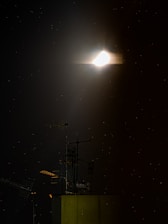 A glowing celestial body is surrounded by a soft halo against a dark night sky. Below it, the silhouette of a building with multiple antennas is visible, highlighting an urban rooftop scene. Tiny stars dot the background, adding to the tranquil ambiance.
