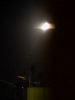 A glowing celestial body is surrounded by a soft halo against a dark night sky. Below it, the silhouette of a building with multiple antennas is visible, highlighting an urban rooftop scene. Tiny stars dot the background, adding to the tranquil ambiance.