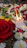 A grouping of lit white candles surrounded by various flowers, including red rose petals, yellow marigolds, and white daisies. Green leaves are interspersed among the red and yellow flower petals, creating a vibrant arrangement on the ground.