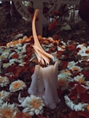 A cluster of white candles is burning amid a bed of flowers, predominantly white chrysanthemums and red roses. The background includes a stone surface with indistinct writing, possibly a memorial or grave marker. The atmosphere is solemn and reverent.