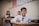 A young student in a white shirt and red tie sits at a wooden desk, holding a pencil poised above an open notebook. Behind him, two other students in similar attire are blurred in the background, suggesting a classroom environment decorated with a colorful chart on the wall.