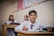 A young student in a white shirt and red tie sits at a wooden desk, holding a pencil poised above an open notebook. Behind him, two other students in similar attire are blurred in the background, suggesting a classroom environment decorated with a colorful chart on the wall.