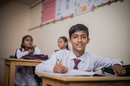 A young student in a white shirt and red tie sits at a wooden desk, holding a pencil poised above an open notebook. Behind him, two other students in similar attire are blurred in the background, suggesting a classroom environment decorated with a colorful chart on the wall.