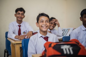 A group of young students in a classroom setting seated at wooden desks. They are dressed in school uniforms consisting of white shirts and red ties. The mood is cheerful, as the students are smiling and appear engaged. Books and other school materials are on the desks.