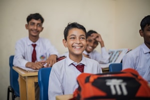 A group of young students in a classroom setting seated at wooden desks. They are dressed in school uniforms consisting of white shirts and red ties. The mood is cheerful, as the students are smiling and appear engaged. Books and other school materials are on the desks.