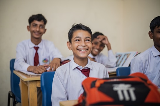 A group of young students in a classroom setting seated at wooden desks. They are dressed in school uniforms consisting of white shirts and red ties. The mood is cheerful, as the students are smiling and appear engaged. Books and other school materials are on the desks.