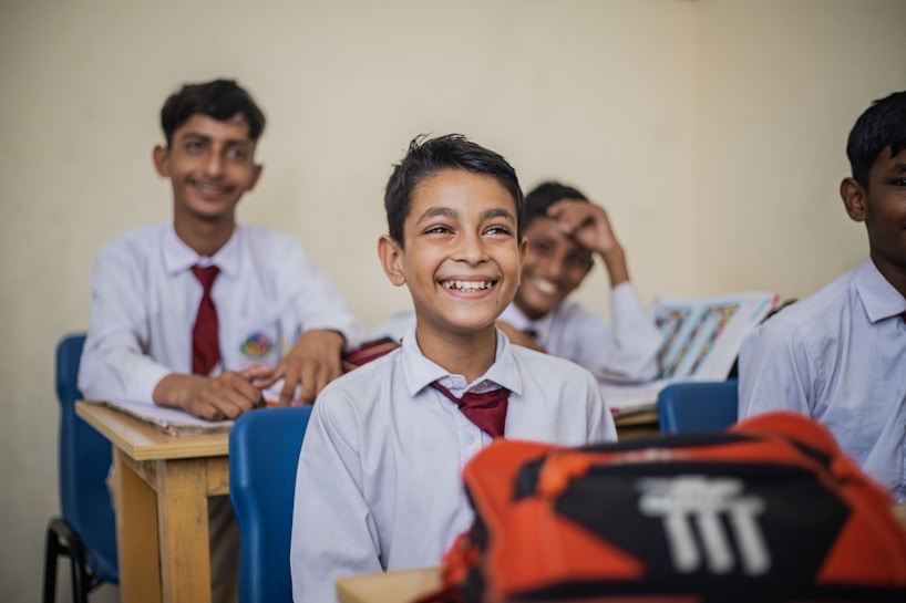 A group of young students in a classroom setting seated at wooden desks. They are dressed in school uniforms consisting of white shirts and red ties. The mood is cheerful, as the students are smiling and appear engaged. Books and other school materials are on the desks.