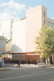 A man in a black suit is walking on the sidewalk next to a parking lot with a large building in the background. The parking lot is mostly empty and partially covered by a roof structure held up by orange beams. There is a large tree with green leaves to the right of the parking lot. The building in the background is modern, with a light pastel façade, and there are additional buildings in the background. The sky is clear with a few clouds.
